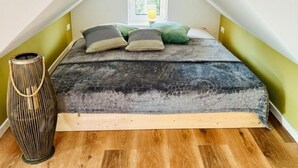 Um quarto com teto inclinado, uma cama com colcha estampada escura, três almofadas, um abajur e um vaso alto com textura em relevo.