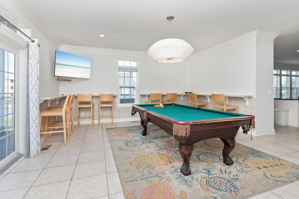 A pool table in a well-lit room with a patterned rug, wooden chairs, and a television on the wall.