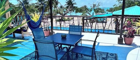A covered outdoor seating area with a table and chairs, a hanging chair, and a view of the beach and ocean.