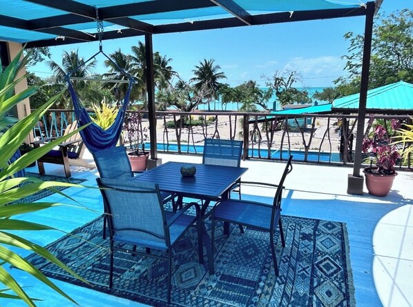 A covered outdoor seating area with a table and chairs, a hanging chair, and a view of the beach and ocean.