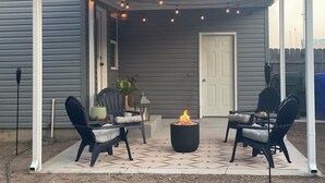 Un patio techado con dos sillas, un pequeño hogar para fuego y luces colgantes.