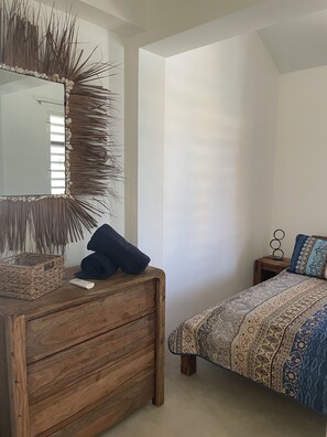 A bedroom with a wooden dresser, a bed with patterned bedding, a mirror with a natural frame, and a small bedside table with a decorative item. - KAS COCOLOCO Lagoon – private waterfront living with resort comfort (Bonaire)