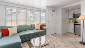 A modern living room with a green sofa, a baby crib, and a kitchen area with a microwave and cabinets.