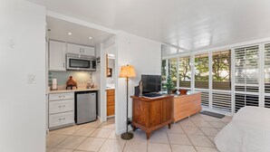 A modern kitchen with white cabinets, a microwave, and a dishwasher. There is a wooden dresser with drawers and a lamp. A bed is visible in the foreground.