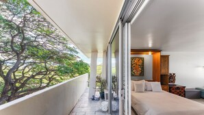A balcony with a view of a tree, a bed with pillows, and a painting on the wall. - Charming Studio Apartment with WiFi, AC in beautiful Diamond Head Beach Park! (Honolulu)