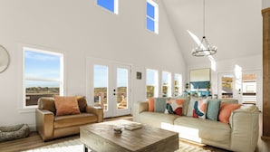 Un salon spacieux avec un plafond haut, de grandes fenêtres, une table basse en bois et un canapé beige agrémenté de coussins colorés.
