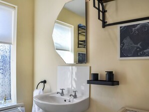 Un baño con un lavabo blanco, dos llaves, un espejo, un perchero para toallas, un estante negro y una ilustración enmarcada de un pez.