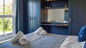 A bedroom with a large window, a bed with a patterned bedspread, two rolled towels, a mirror, and a dark blue wardrobe with shelves.