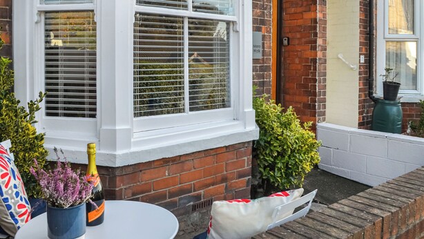 A corner of a building with a white window, a brick wall, and a small garden area with a table, a bottle, and a bag.