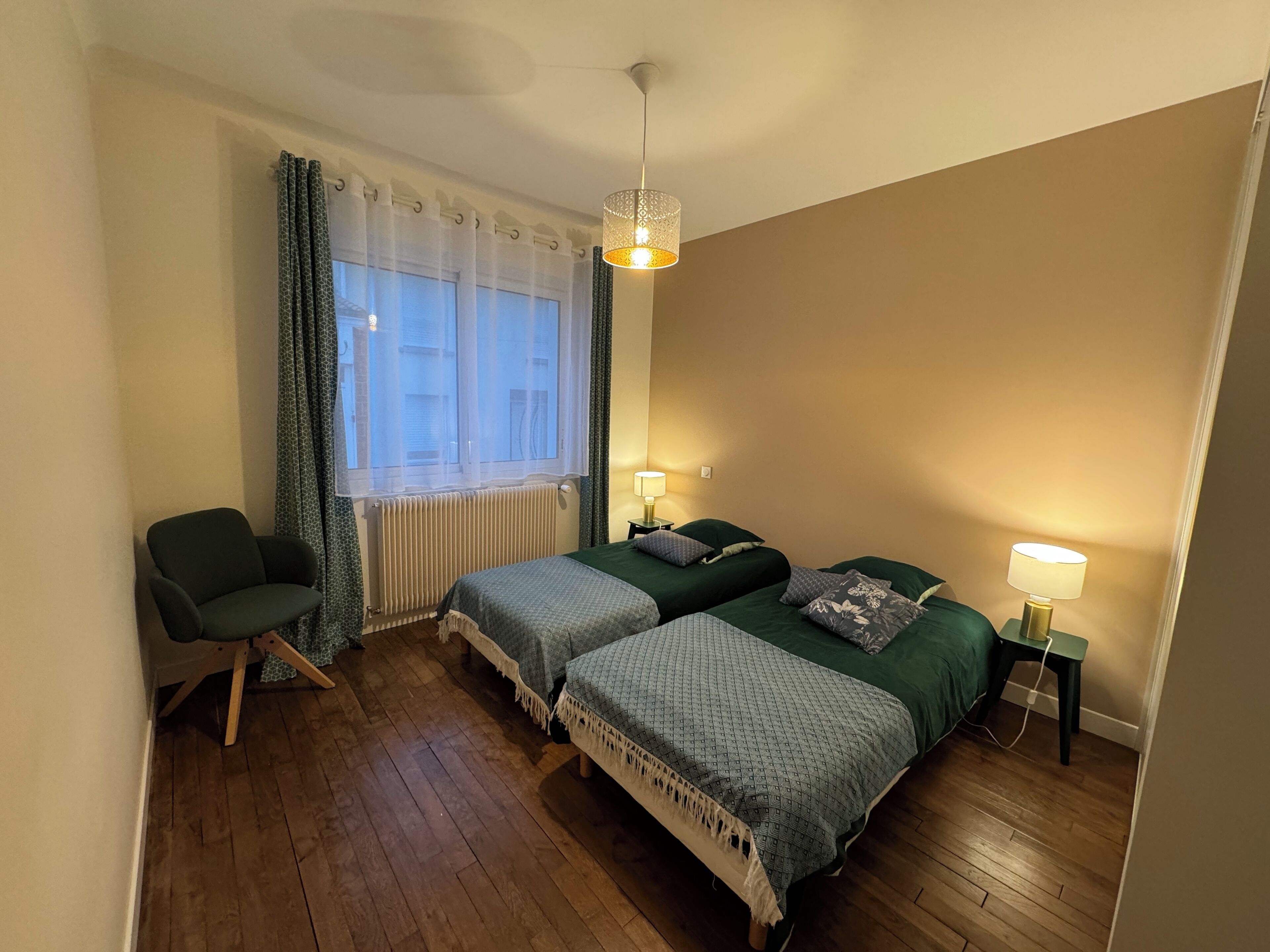 Une chambre avec deux lits, une chaise, une fenêtre avec des rideaux et un luminaire suspendu.
