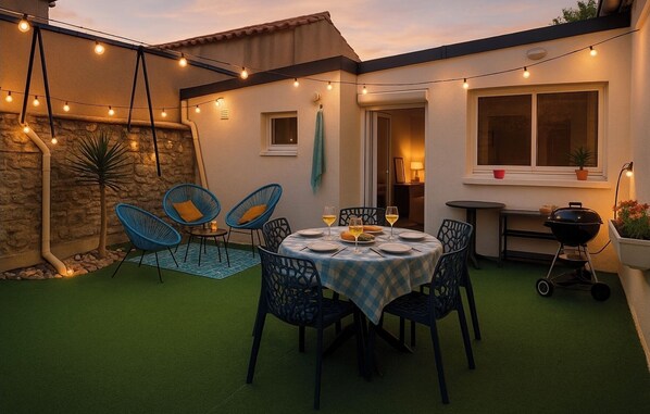 Une terrasse extérieure avec une table ronde dressée pour le repas, entourée de chaises bleues, et des guirlandes lumineuses suspendues au-dessus.