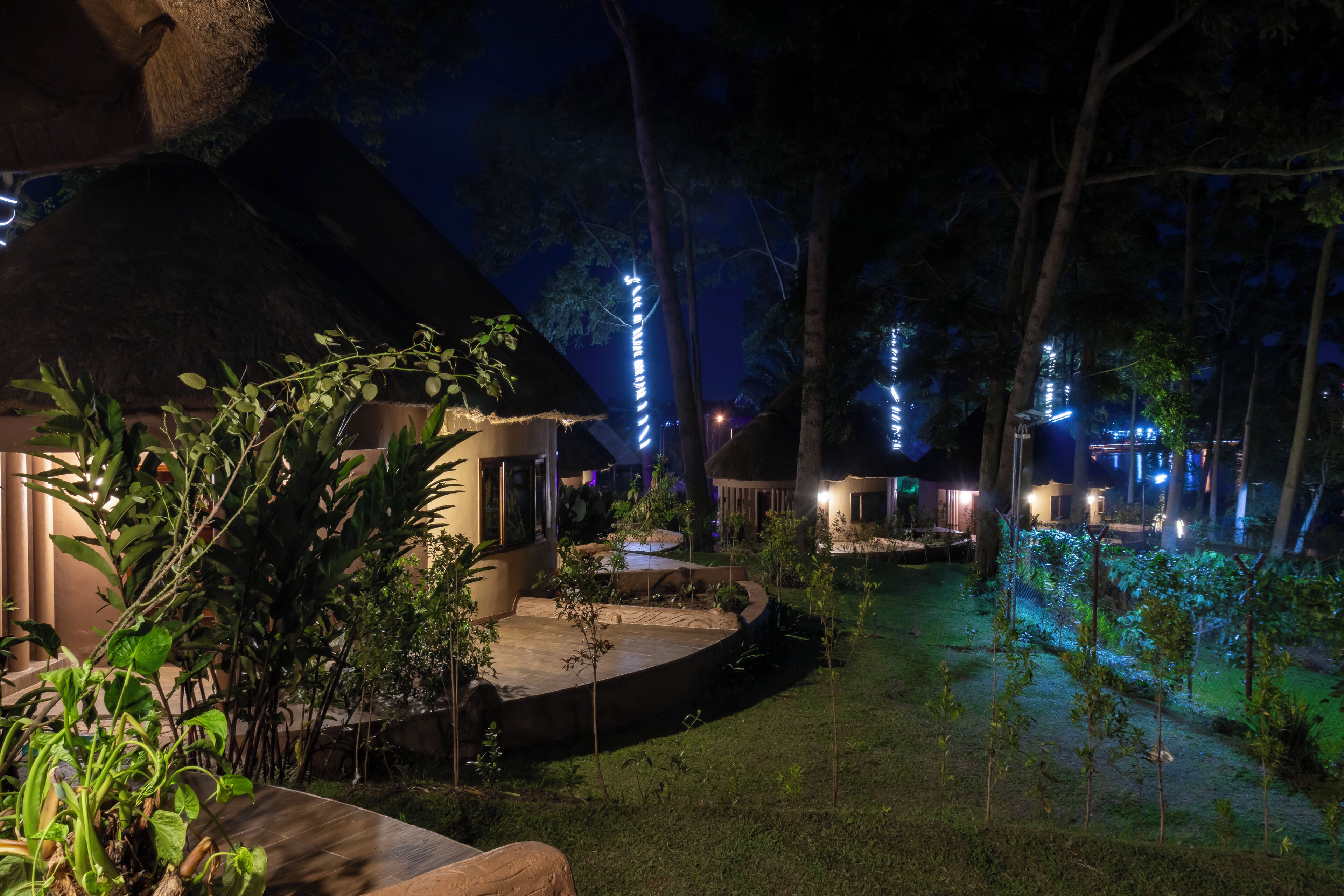 A well-lit outdoor area with thatched-roof structures, surrounded by greenery and illuminated string lights.