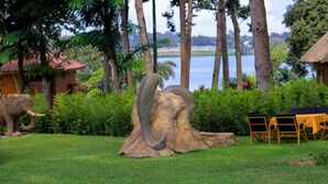 A sculpture of a large elephant in a grassy area with a table and chairs, surrounded by trees and a body of water in the background. - De Rain Eco Beach Resort (Entebbe)