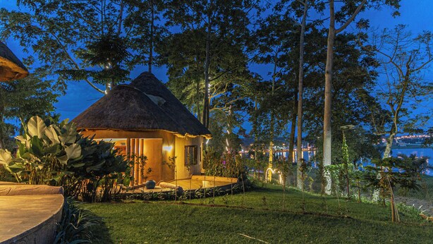 A thatched-roof house with a well-lit patio, surrounded by lush greenery and tall trees. - De Rain Eco Beach Resort (Entebbe)