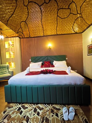 A hotel room with a large bed, a green upholstered headboard, a patterned rug, and a pair of slippers on the floor.