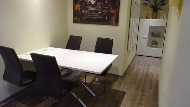 Un coin repas avec une table blanche et quatre chaises noires, un tableau au mur et un meuble blanc orné d’une plante.
