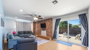 Un salon moderne avec un mur en briques, des canapés gris, un ventilateur de plafond et une porte-fenêtre coulissante donnant sur un jardin.