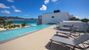 Une piscine sur le toit, agrémentée de chaises longues, offrant une vue dégagée sur l’océan et s’inscrivant dans un décor composé d’un bâtiment moderne en arrière-plan.