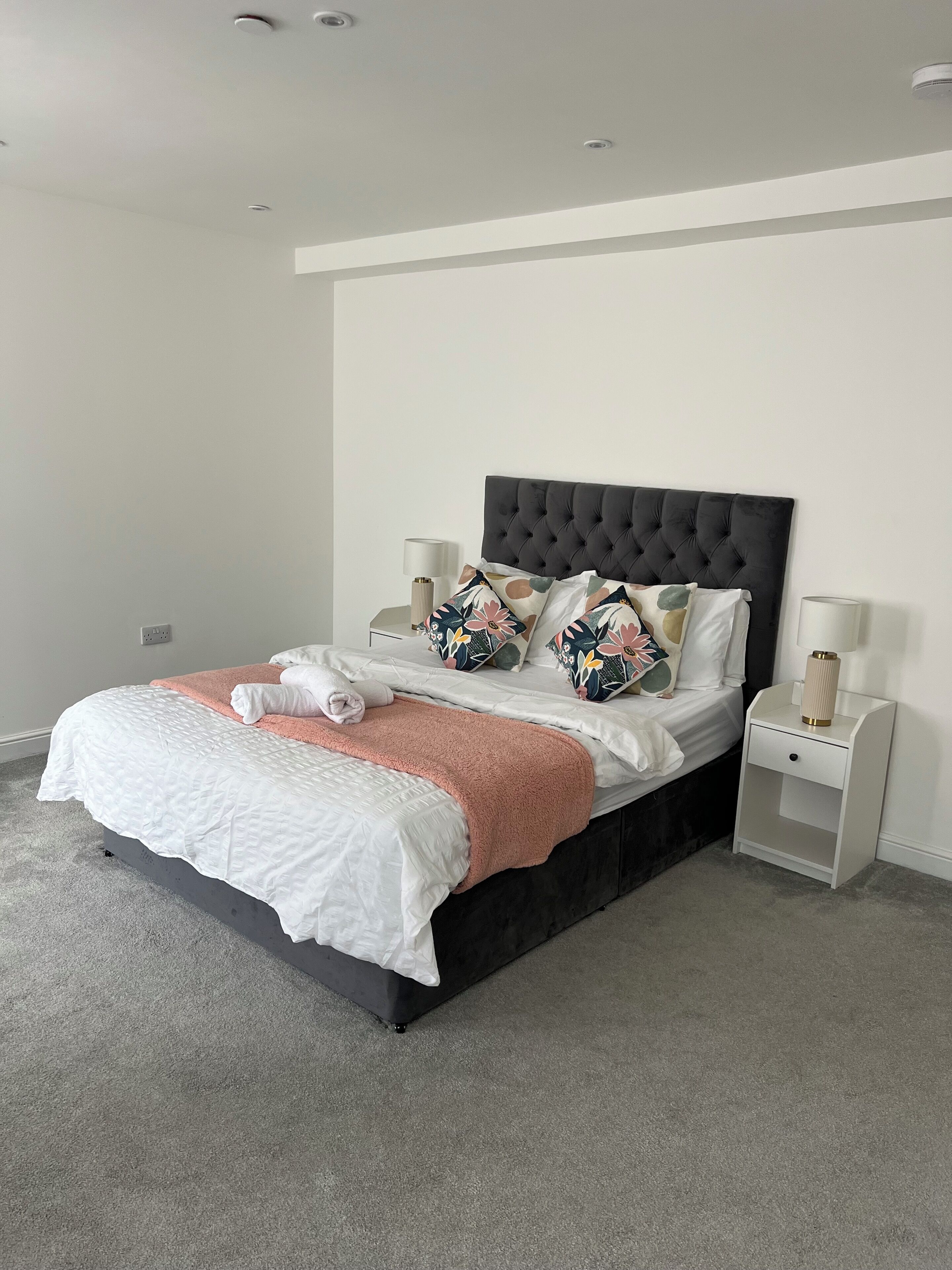 A neatly made bed with a white duvet, a peach throw blanket, and floral-patterned pillows. There are two bedside tables with lamps, and the room has a modern, minimalist design with a grey carpet.