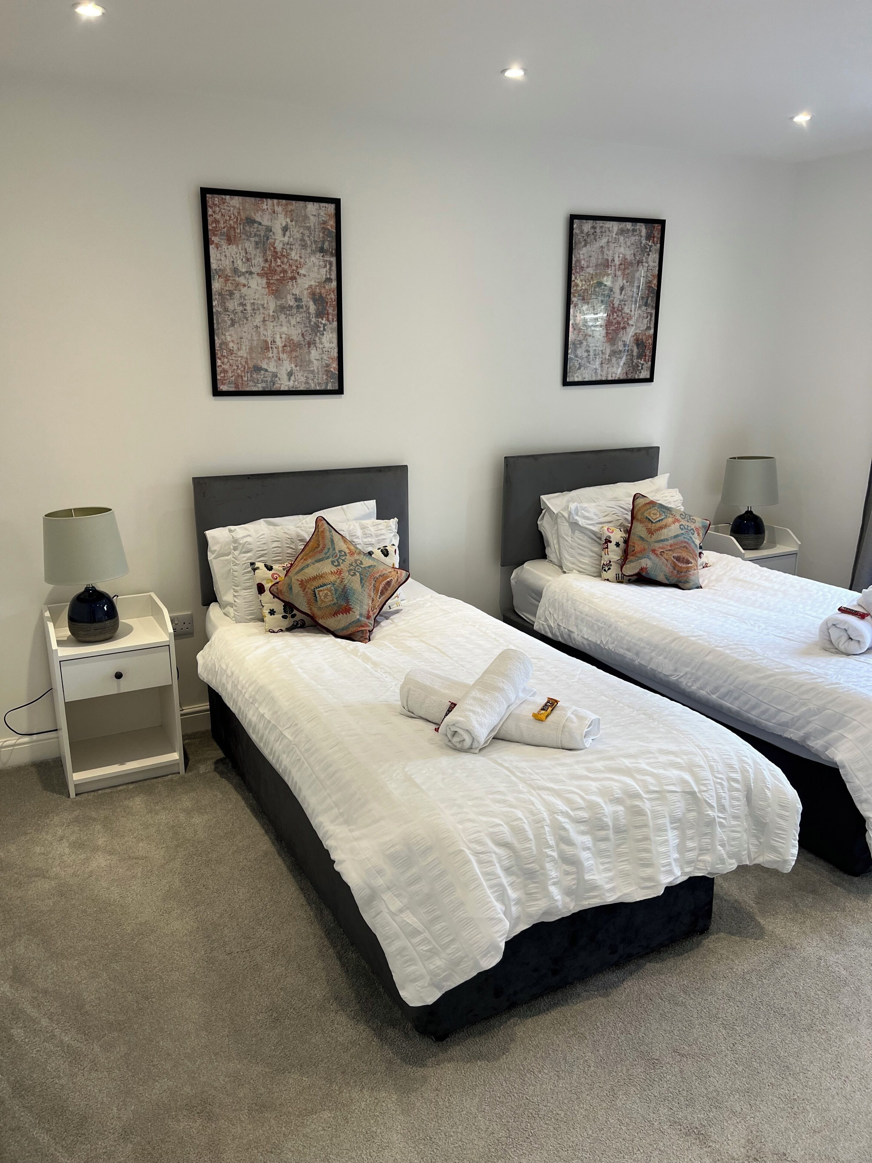 Une chambre d’hôtel avec deux lits, du linge de lit blanc, des coussins décoratifs, des lampes de chevet et des œuvres d’art abstraites accrochées aux murs.