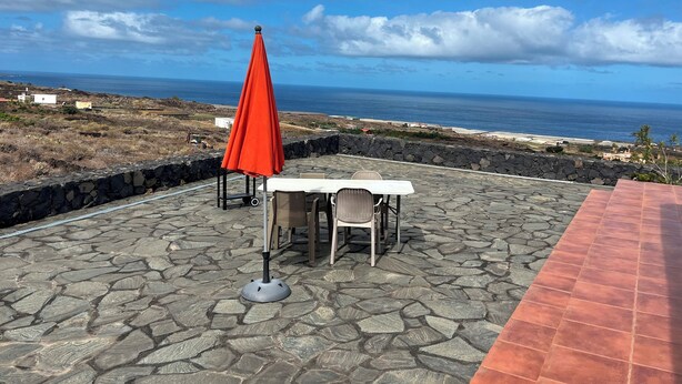 Un patio exterior con una mesa y sillas, una sombrilla roja y vistas al océano.