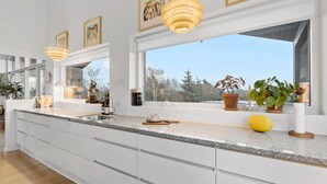 A modern kitchen with a large island, white cabinetry, and a view of trees through large windows. - 3 bedroom beautiful home in Nibe (Nibe)