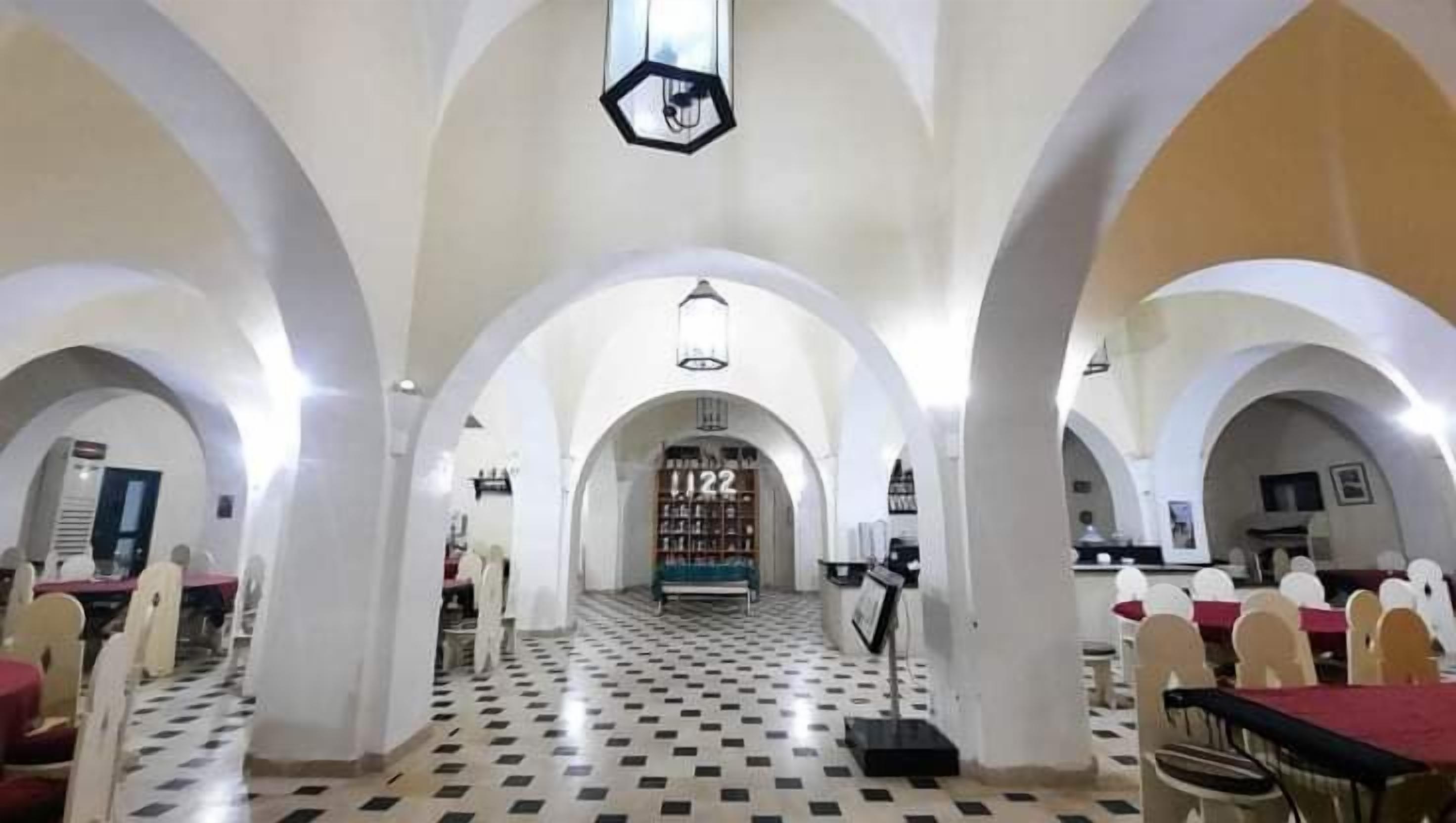 A spacious hall with high ceilings and arched alcoves, a checkered floor, and rows of tables and chairs.