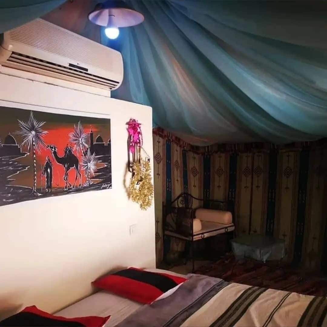 A room with a bed, a wall-mounted air conditioner, a painting of camels, and a hanging decoration.