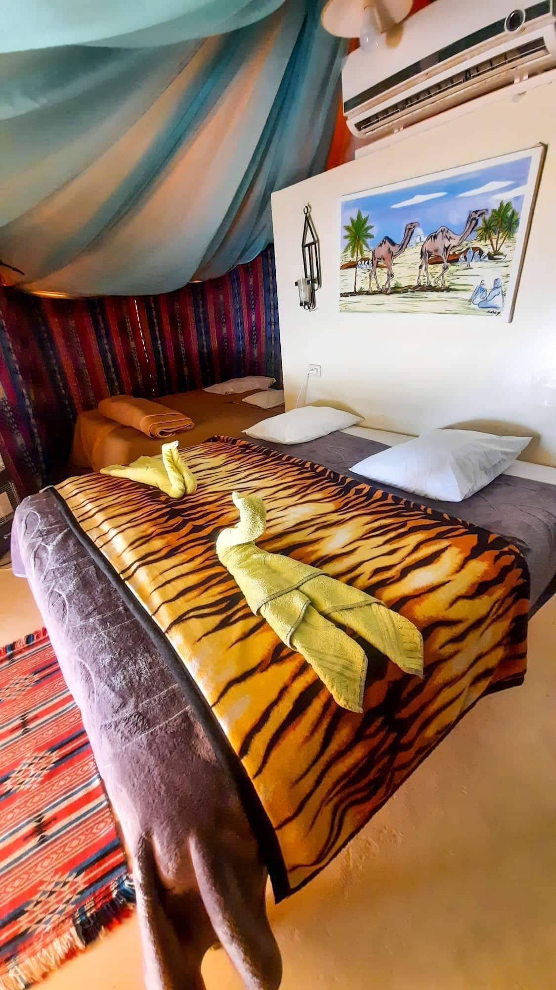 A bed with a tiger print bedspread, a wall-mounted air conditioner, and a painting of a desert scene.