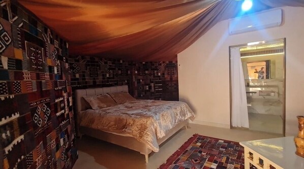 A bedroom with a bed, a patterned wall, a small table, and a door leading to a bathroom. - Hotel Pansy (Douz Sud)