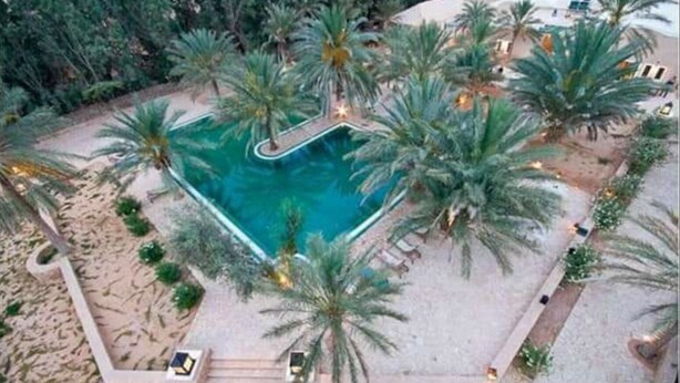 Vue aérienne d’un complexe hôtelier avec une piscine centrale entourée de palmiers et d’une zone sablonneuse.