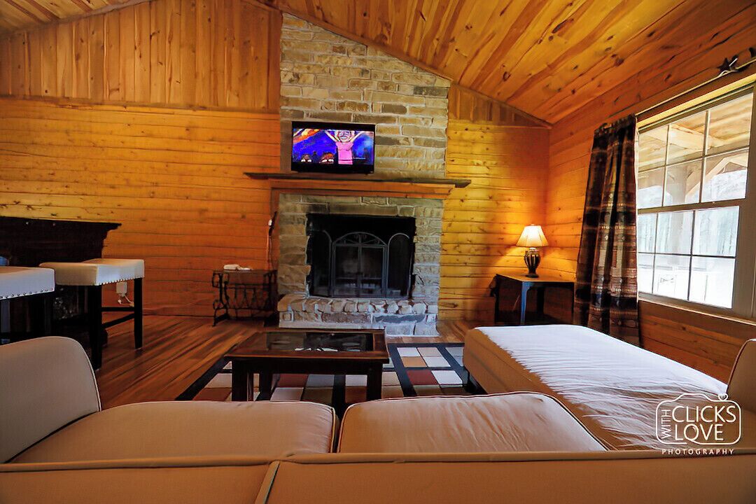 A cozy cabin interior with a stone fireplace, wooden walls, a television mounted above it, and a bed with a view of the outside.