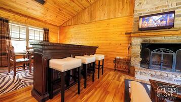 A cozy room with wooden walls and a stone fireplace. There are three bar stools with cushioned seats and a wooden bar. A television is mounted above the fireplace.