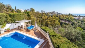 Un espace piscine agrémenté de chaises longues et d’une pergola, entouré d’une végétation luxuriante et offrant une vue sur une ville côtière.