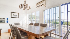 Un espace repas avec une grande table en bois, des chaises à revêtement gris, un lustre et de grandes fenêtres donnant sur un paysage verdoyant et des bâtiments.