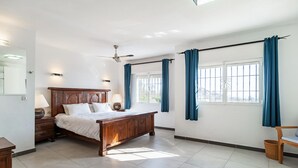 Une chambre à coucher avec un lit en bois, une table de chevet, une lampe, un ventilateur de plafond et une fenêtre avec des rideaux.
