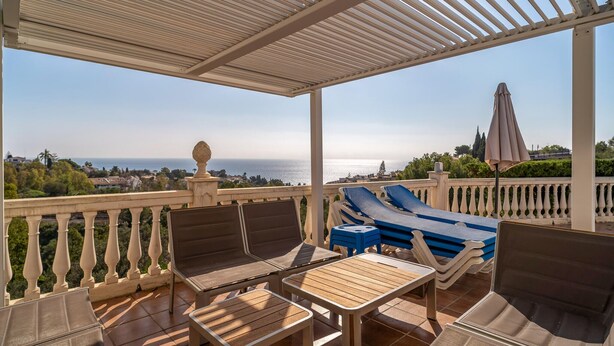 Un espace extérieur aménagé, abrité et doté de chaises longues et de tables, offrant une vue panoramique sur la mer et une ville côtière.