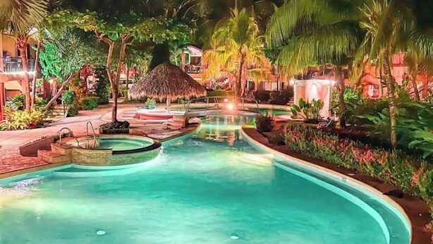 Een zwembadterras van een resort bij nacht, met een helder zwembad, palmbomen en constructies met rieten daken.