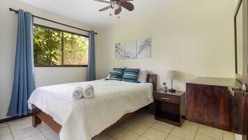 A bedroom with a bed, a nightstand, a ceiling fan, a window with curtains, and a wall with artwork.