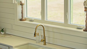 Eine Küche mit einem großen Fenster mit Blick auf einen Strand, einem Waschbecken mit goldenem Wasserhahn und weißen Schränken.