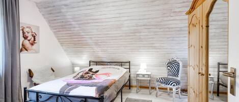 Un dormitorio con techo inclinado, una cama con manta colorida, una silla con estampado de cebra, un armario de madera y una mesita con lámpara.