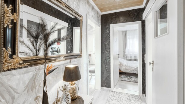 A hallway with a marble wall, a large mirror, a lamp, and a door leading to a bedroom.