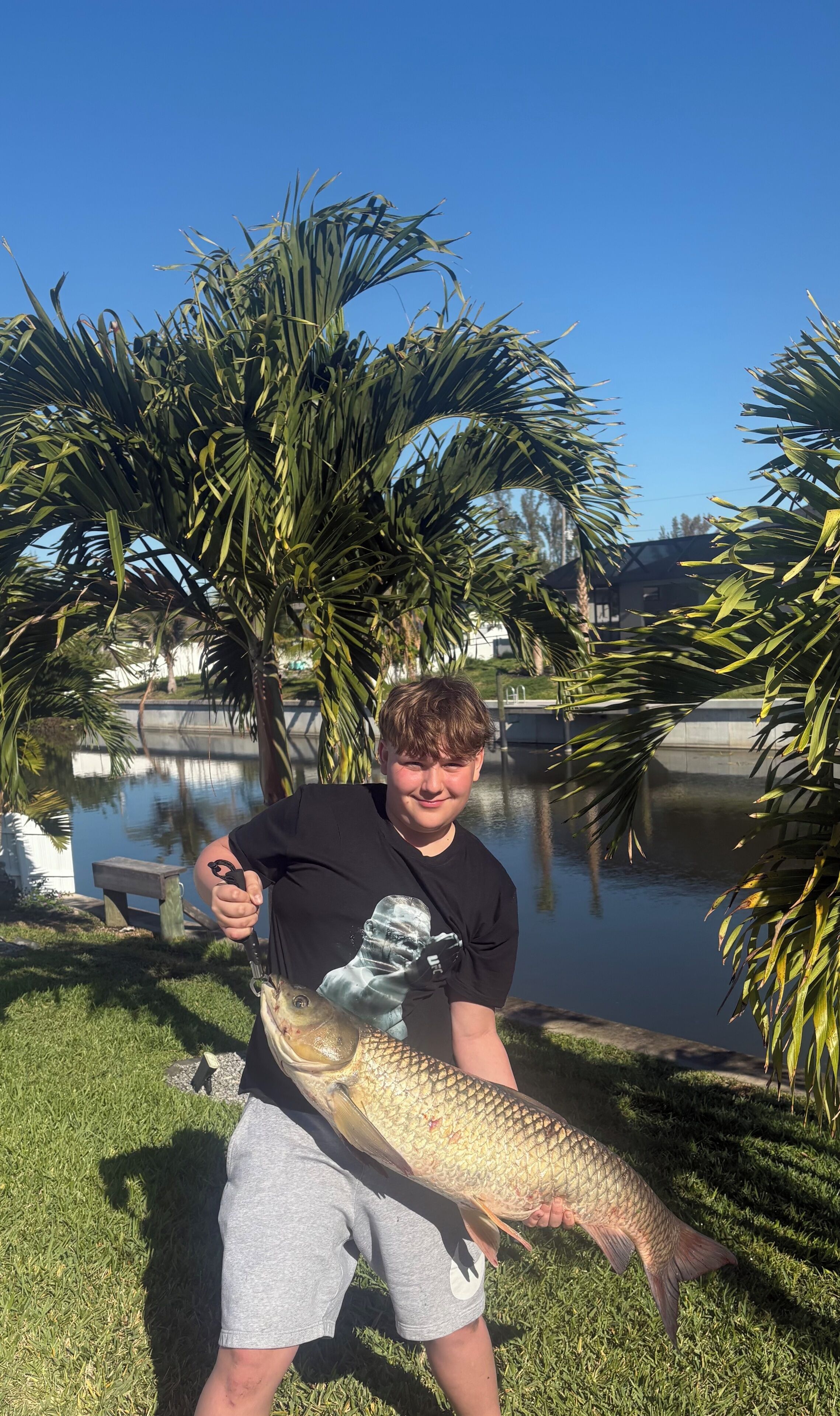 A person holding a large fish by a water body with palm trees and a building in the background.
