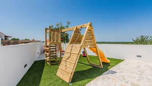 Una estructura de juego de madera con un tobogán y elementos para escalar, sobre césped artificial y un patio de piedra al frente.