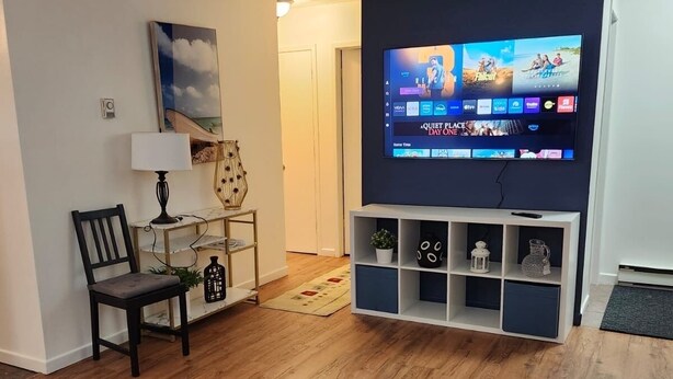Una sala de estar con un televisor montado en la pared, un sofá, una silla y una mesita de noche con una lámpara.