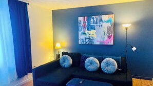 Una sala de estar moderna con un sofá seccional, dos almohadones decorativos, un pequeño reposapiés, una lámpara de pie y una obra de arte abstracta en la pared.