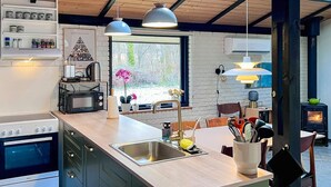 A modern kitchen with a central island, built-in oven, microwave, and a stove. There is a window providing a view of the outdoors, and a wood-burning stove in the dining area. - 4 Person Holiday Home in Farvang (Farvang)