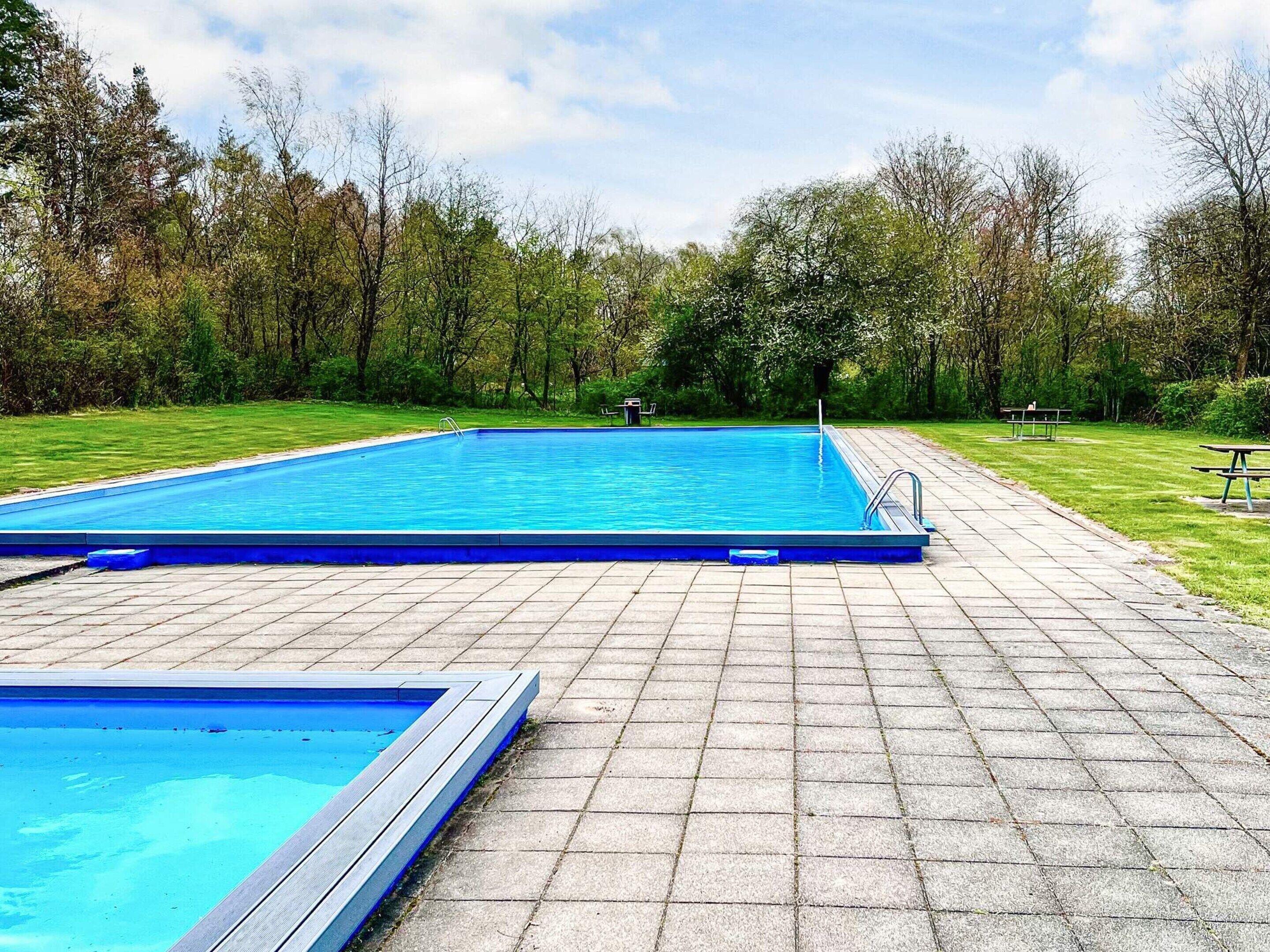A rectangular swimming pool with a tiled deck, surrounded by a grassy area and trees.