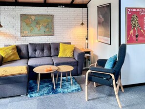 A modern living room with a grey sofa, two round wooden tables, a blue armchair, a world map on the wall, and a poster of a giraffe. - 4 Person Holiday Home in Farvang (Farvang)