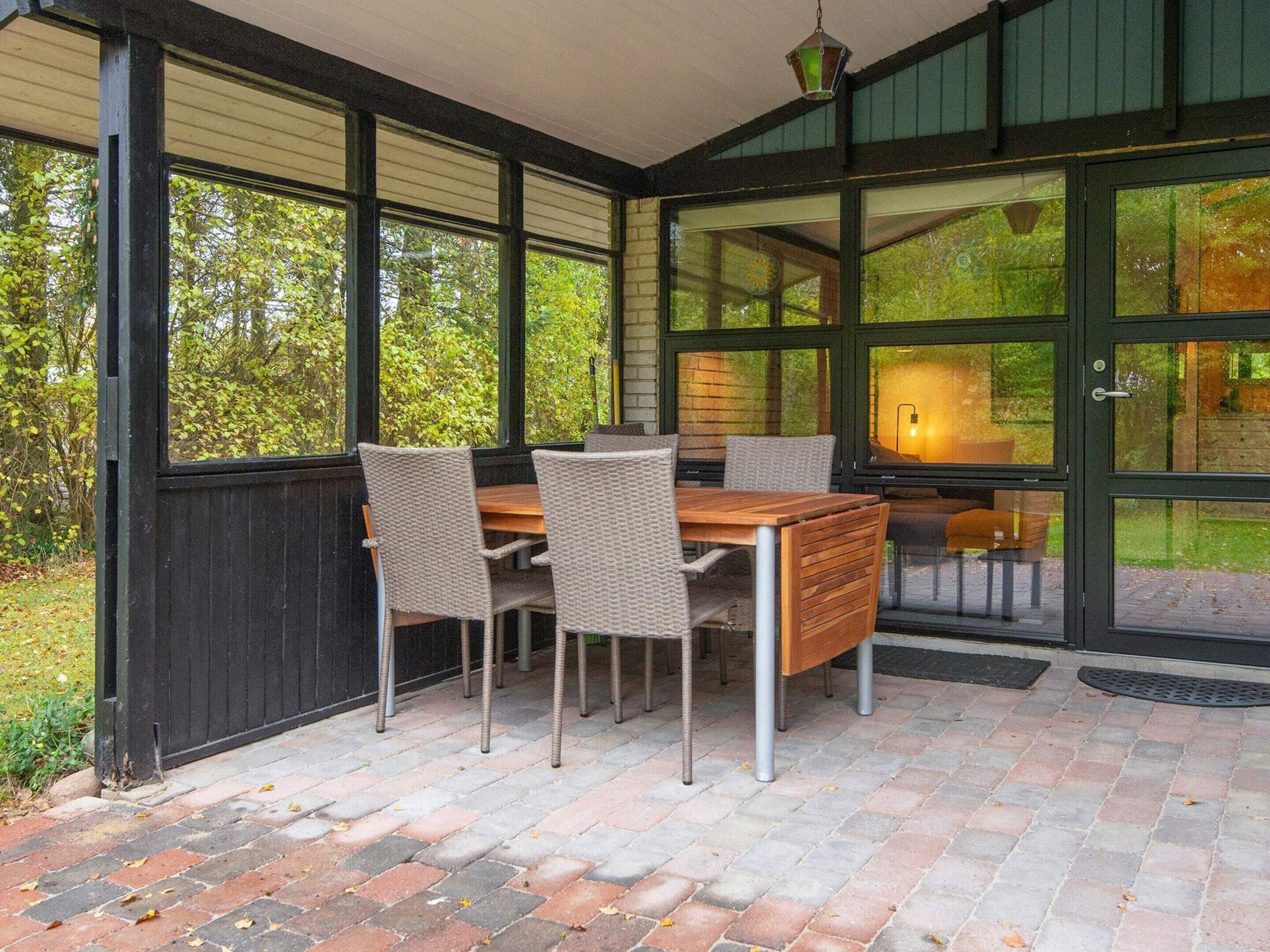 A covered patio with a table and chairs, large windows, and a view of greenery.